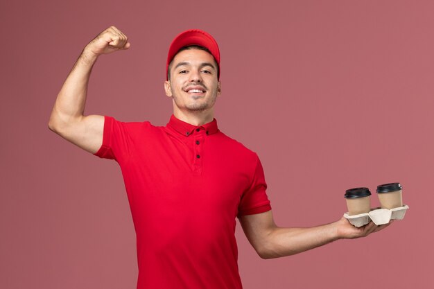 Курьер-мужчина, вид спереди в красной форме, держит коричневые кофейные чашки и сгибает розовую стену.