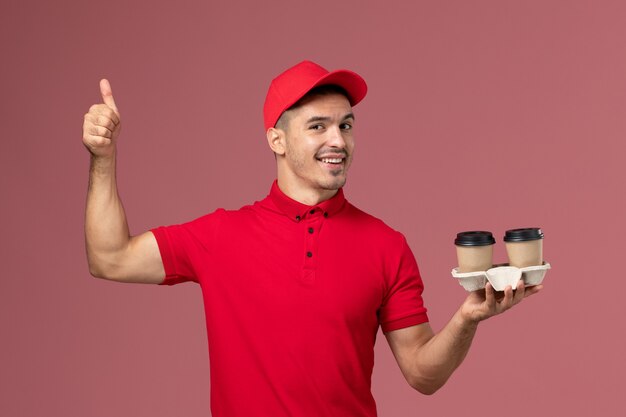 Курьер-мужчина в красной униформе, держащий коричневые кофейные чашки на светло-розовой стене, рабочий