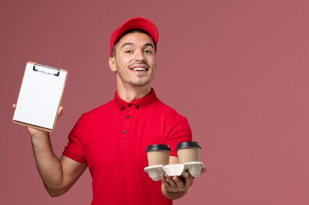 Front view male courier in red uniform holding brown delivery coffee cups and notepad with smile on pink wall worker