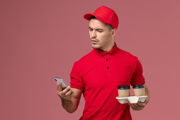 Front view male courier in red uniform holding brown delivery coffee cups and phone on the light-pink desk  worker
