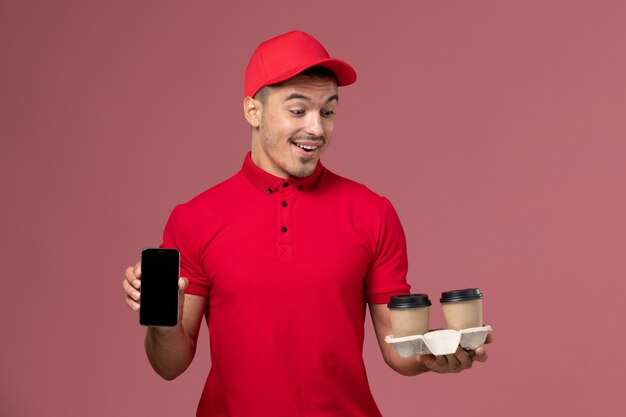 Front view male courier in red uniform holding brown delivery coffee cups and phone on the light-pink wall worker job