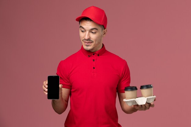 Front view male courier in red uniform holding brown delivery coffee cups and phone on the light-pink wall worker male
