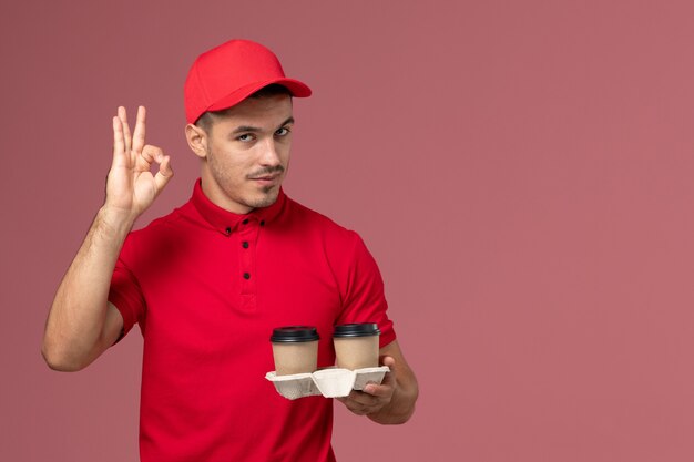 Front view male courier in red uniform holding brown delivery coffee cups on pink desk  worker