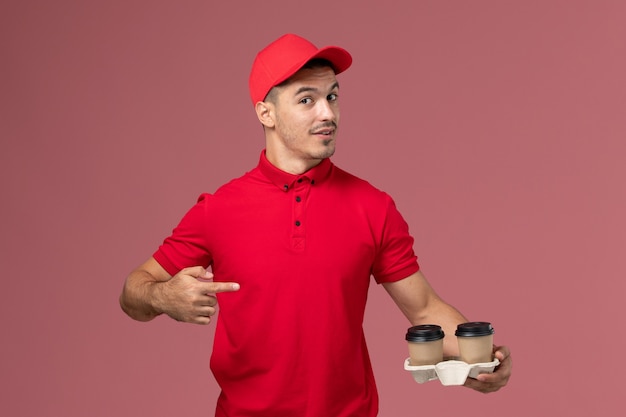 Front view male courier in red uniform holding brown delivery coffee cups on the pink desk 