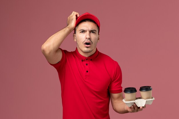 Front view male courier in red uniform holding brown delivery coffee cups on the pink wall  male worker