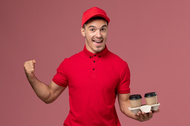 Front view male courier in red uniform holding brown delivery coffee cups on the pink wall 