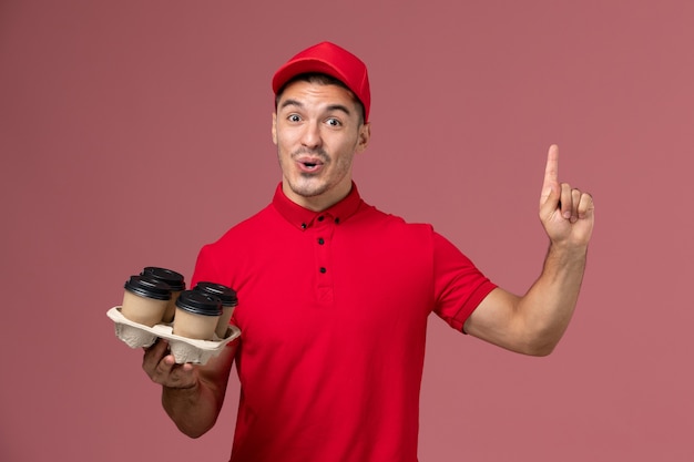Front view male courier in red uniform holding brown delivery coffee cups on the pink wall 