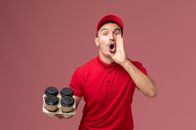 Front view male courier in red uniform holding brown delivery coffee cups screaming on light pink wall worker