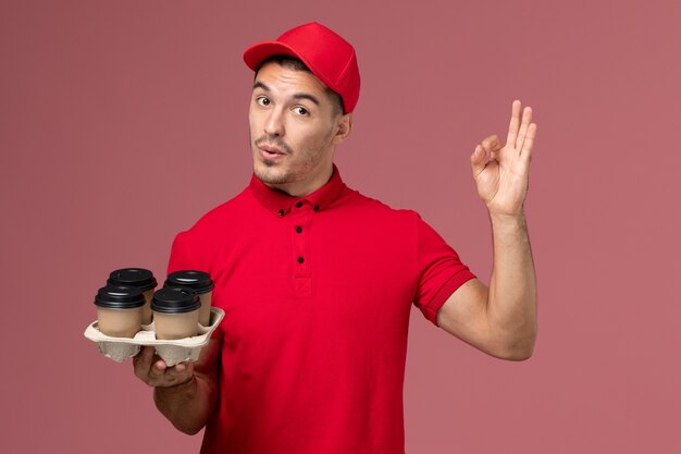 Front view male courier in red uniform holding brown delivery coffee cups showign alright sign on pink wall 