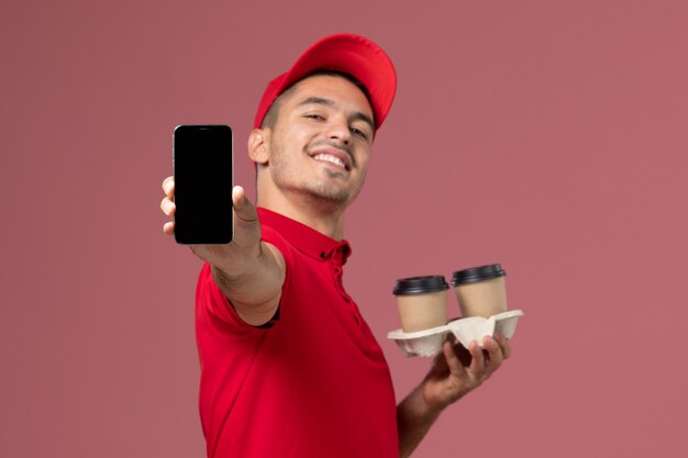 Front view male courier in red uniform holding brown delivery coffee cups showing phone on the pink wall 