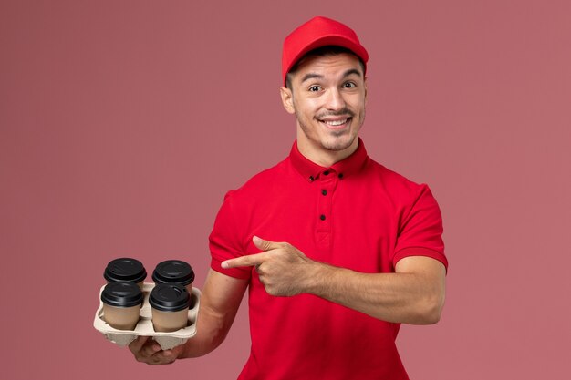 Front view male courier in red uniform holding brown delivery coffee cups and smiling on pink wall worker