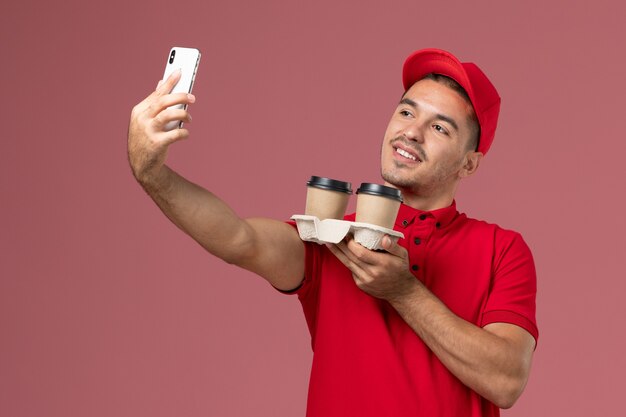 Front view male courier in red uniform holding brown delivery coffee cups taking a selfie with them on pink wall 