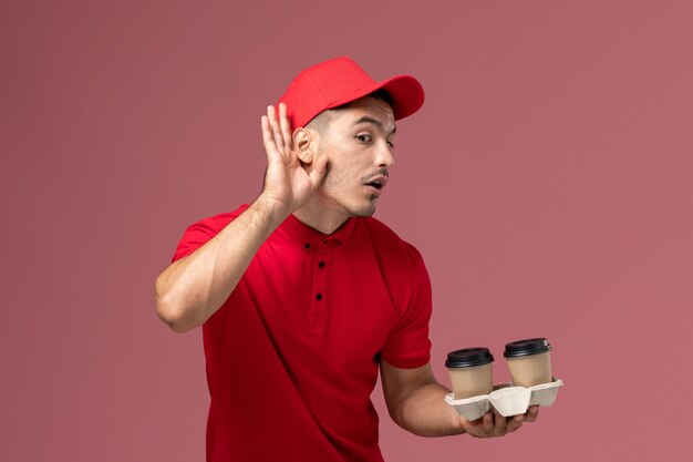 Front view male courier in red uniform holding brown delivery coffee cups trying to hear out on the pink wall 