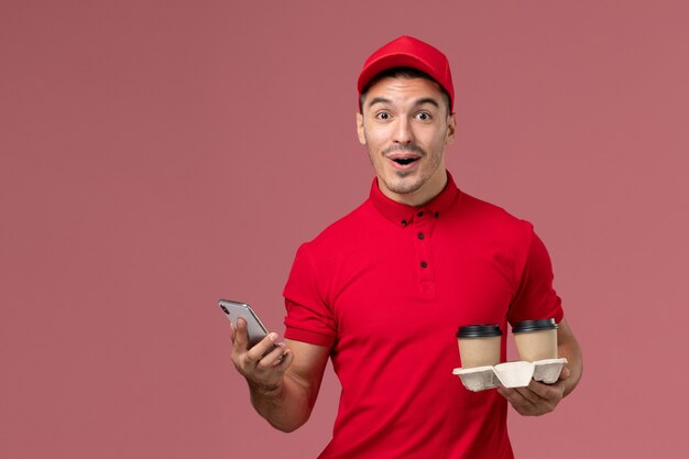 Front view male courier in red uniform holding brown delivery coffee cups and using a phone on the light pink wall 