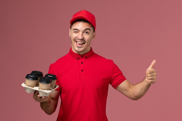 Front view male courier in red uniform holding brown delivery coffee cups winking on pink wall 