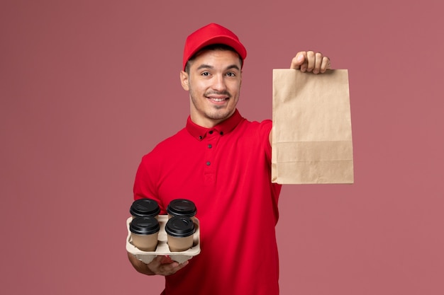 ピンクの壁に食品パッケージと茶色の配達コーヒーカップを保持している赤い制服の正面図男性宅配便サービス配達労働者の制服