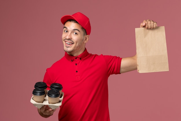 Front view male courier in red uniform holding brown delivery coffee cups with food package on the pink wall 