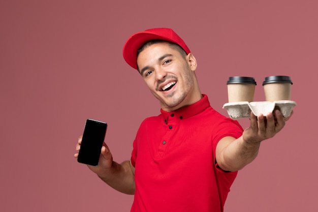 Front view male courier in red uniform holding brown delivery coffee cups with phone on pink wall 