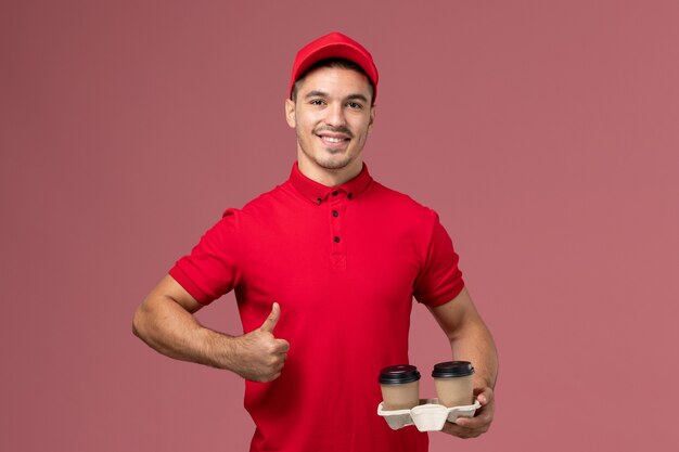 Front view male courier in red uniform holding brown delivery coffee cups with slight smile on pink wall  worker male