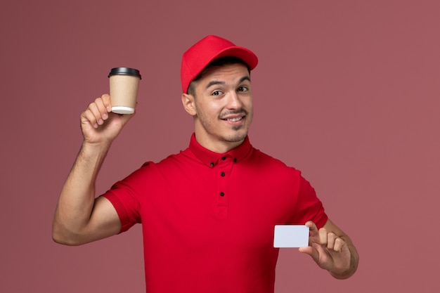 Курьер-мужчина, вид спереди в красной форме, держит чашку кофе с доставкой и открытку на розовой стене
