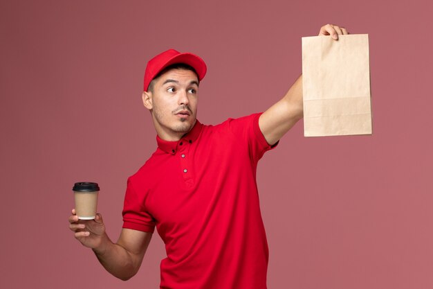 Front view male courier in red uniform holding delivery coffee cup and food package on light pink wall service delivery worker male uniform job