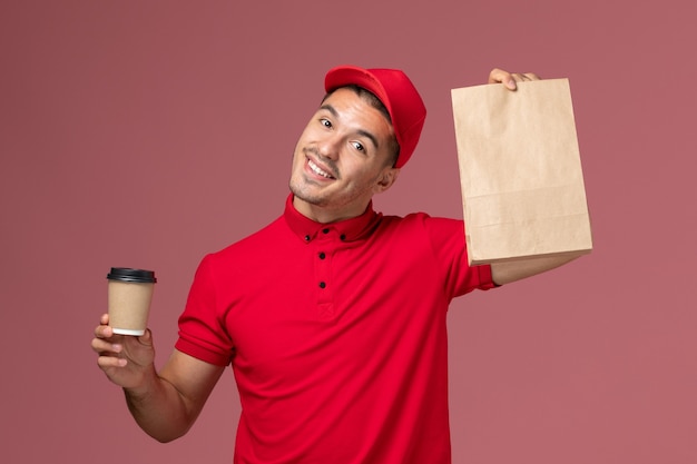 Corriere maschio di vista frontale in uniforme rossa che tiene la tazza di caffè di consegna e il pacchetto alimentare sul lavoro uniforme dell'operaio di consegna di servizio della parete rosa