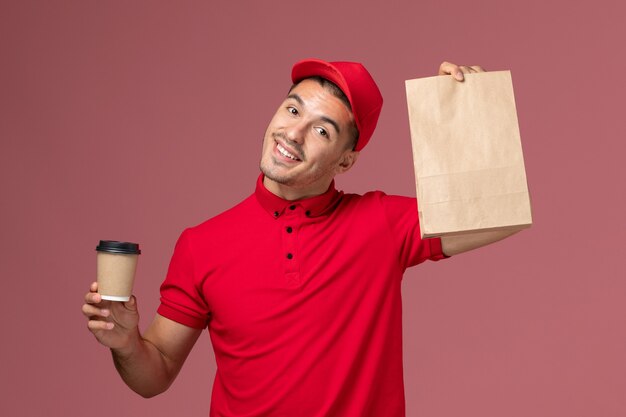Front view male courier in red uniform holding delivery coffee cup and food package on pink wall service delivery worker uniform job