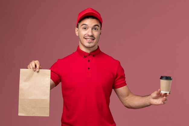 Front view male courier in red uniform holding delivery coffee cup and food package on the pink wall 