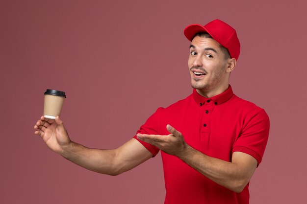 淡いピンクの壁の男性労働者に配達コーヒーカップを保持している赤い制服の正面図男性宅配便