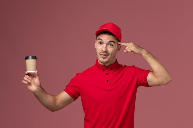 Front view male courier in red uniform holding delivery coffee cup on light-pink wall 