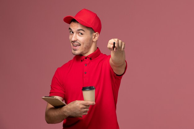 Front view male courier in red uniform holding delivery coffee cup and notepad on pink floor  worker male work