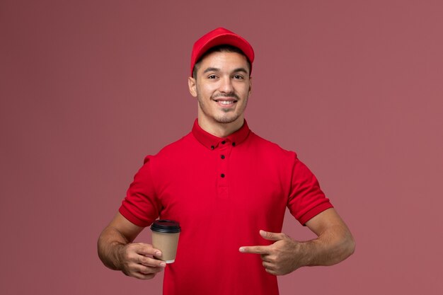 Front view male courier in red uniform holding delivery coffee cup on pink wall job male worker