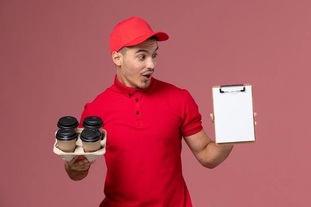 Курьер-мужчина в красной форме, вид спереди, держит в руках кофейные чашки с блокнотом на розовой стене рабочего
