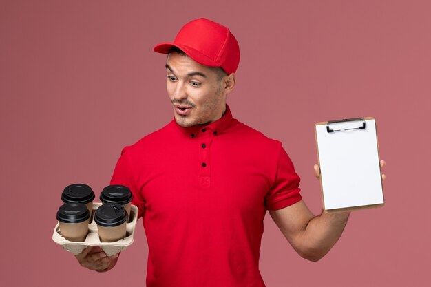 ピンクの壁にメモ帳と配達コーヒーカップを保持している赤い制服の正面図男性宅配便