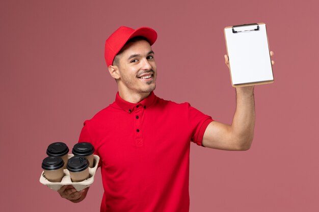 ピンクの壁にメモ帳の笑顔で配達コーヒーカップを保持している赤い制服の正面図男性宅配便
