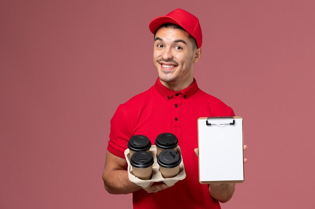 Front view male courier in red uniform holding delivery coffee cups with notepad smile on pink wall 