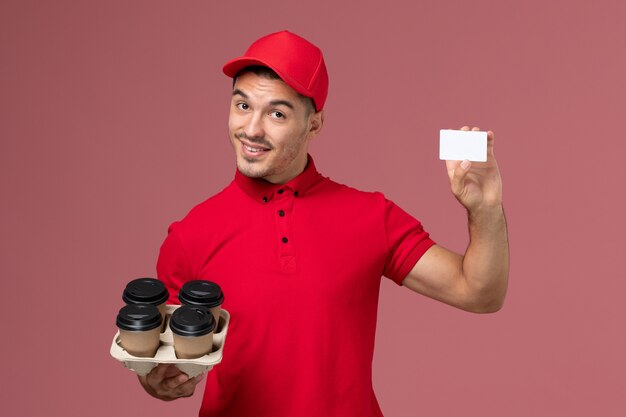 분홍색 벽 서비스 배달 작업 작업자 유니폼에 흰색 카드와 함께 배달 커피 컵을 들고 빨간색 유니폼에 전면보기 남성 택배
