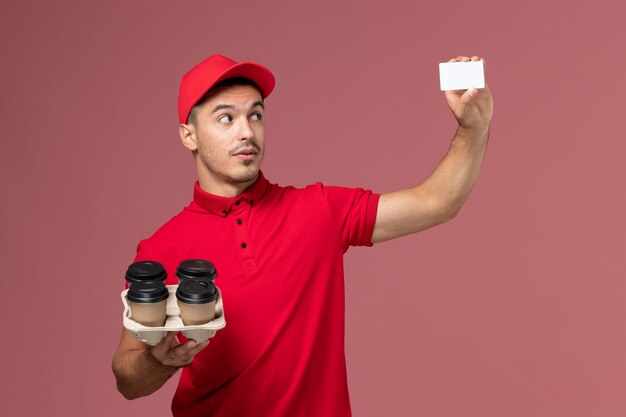 Front view male courier in red uniform holding delivery coffee cups with white card on the pink wall service delivery job worker uniform