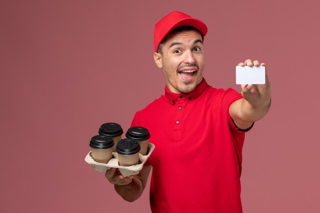 Front view male courier in red uniform holding delivery coffee cups with white card on the pink wall service job delivery worker uniform