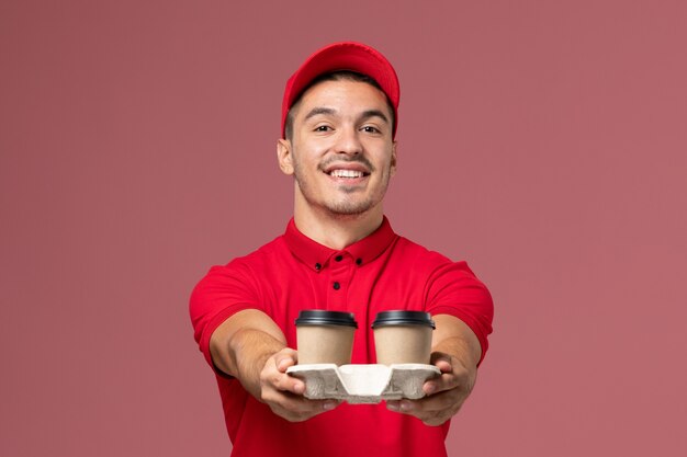 Corriere maschio di vista frontale in uniforme rossa che sorride e che tiene le tazze di caffè marroni di consegna sulla parete rosa