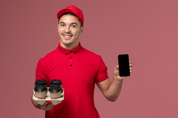 Front view male courier in red uniform smiling and holding delivery coffee cups with phone on pink wall 