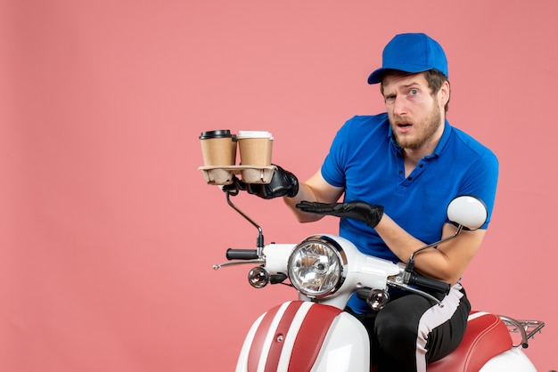 Front view male courier sitting on bike and holding coffee cups on a pink 