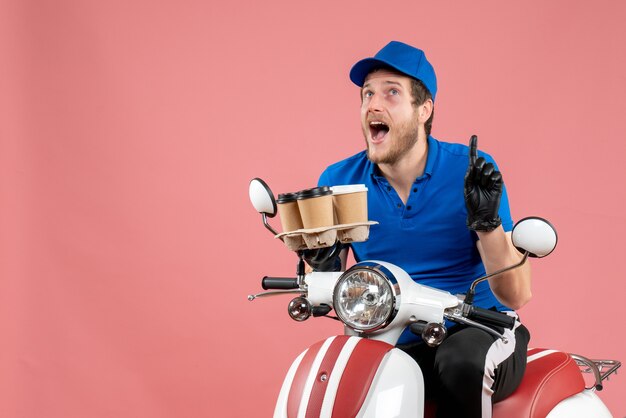 Front view male courier sitting on bike and holding coffee cups on a pink 