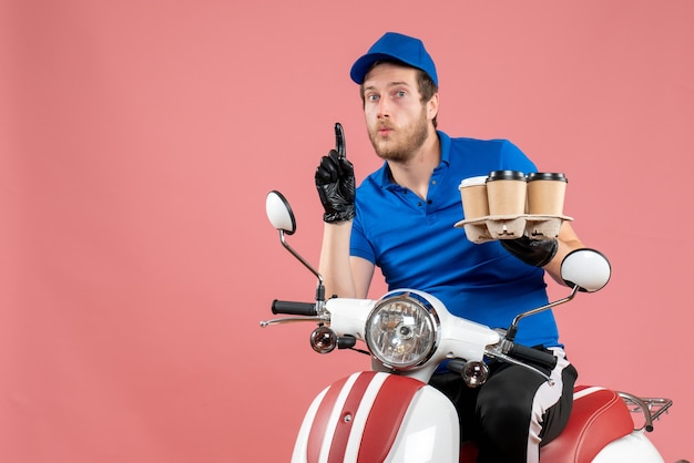 Front view male courier sitting on bike and holding coffee cups on a pink 