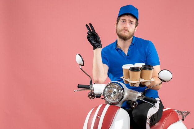 Front view male courier sitting on bike and holding coffee cups on a pink 