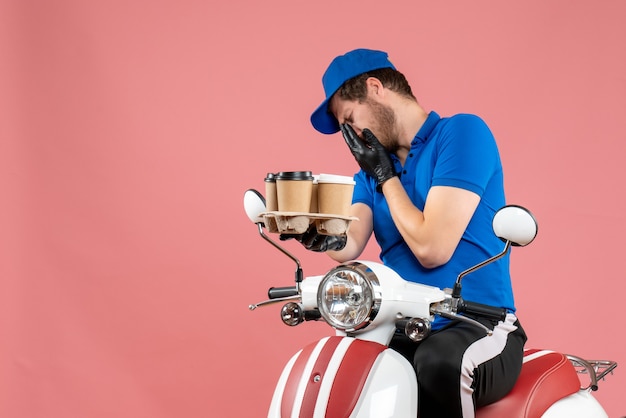 Front view male courier sitting on bike and holding coffee cups on pink 