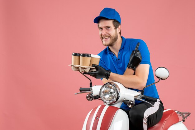 Front view male courier sitting on bike and holding coffee cups on pink 