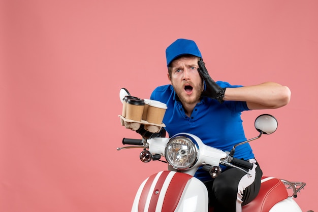 Front view male courier sitting on bike and holding coffee cups on pink 