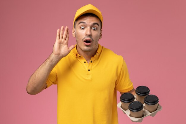 Front view male courier in yellow uniform calling out and holding delivery coffee cups on the pink desk 