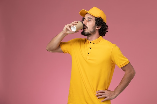 노란색 유니폼과 케이프 핑크 벽에 커피를 마시는 전면보기 남성 택배
